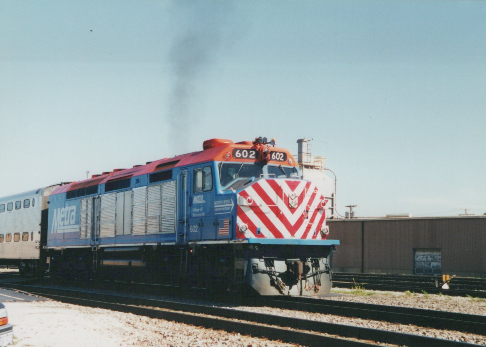 A blue Metra diesel locomotive with the number 602 and red-and-white striped nose is on tracks in a yard on a clear day, pulling a stainless steel passenger car.