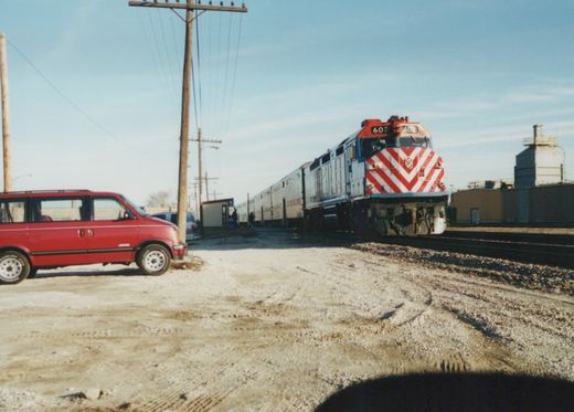 A red, white, and blue Metra locomotive numbered 602 stands on railroad tracks next to a red minivan in a gravel yard under a clear winter sky.
