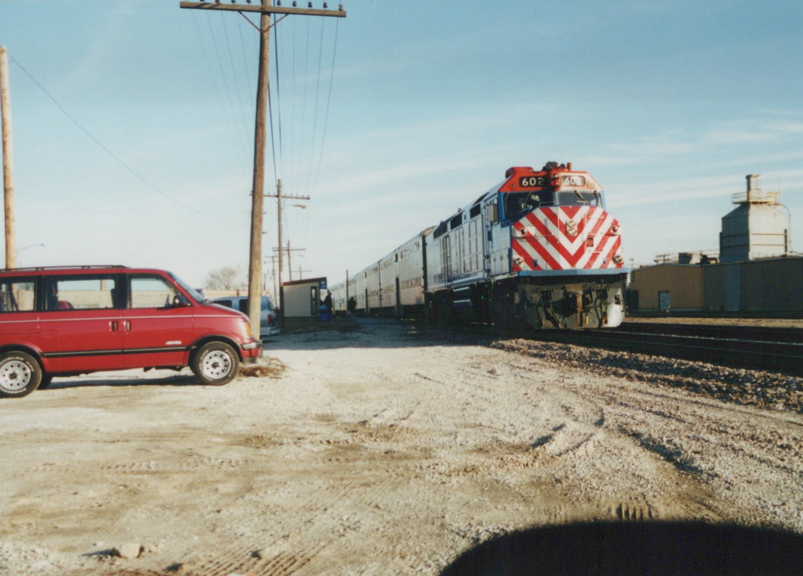 A red, white, and blue Metra locomotive numbered 602 stands on railroad tracks next to a red minivan in a gravel yard under a clear winter sky.