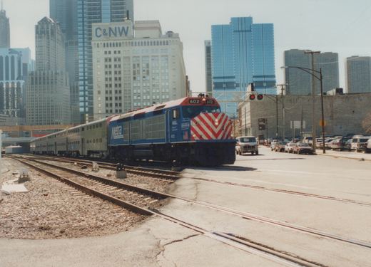 A blue and red Metra locomotive numbered 602 leads a train of silver double-deck passenger cars across tracks in a city street, with tall buildings behind it.