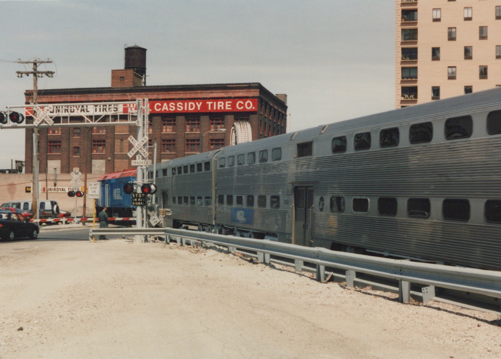A silver Metra gallery car with the number 7284 is stopped at a grade crossing, with a Uniroyal Tires building in the background and a person standing nearby.
