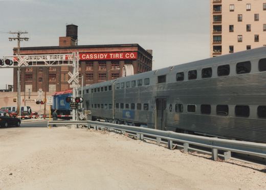 A silver Metra gallery car with the number 7284 is stopped at a grade crossing, with a Uniroyal Tires building in the background and a person standing nearby.