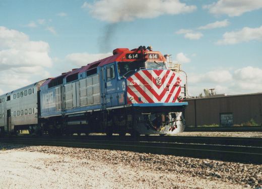 A blue and red Metra locomotive numbered 614, with a white and red chevron front, pulls a silver passenger car on a railroad track under a partly cloudy sky.