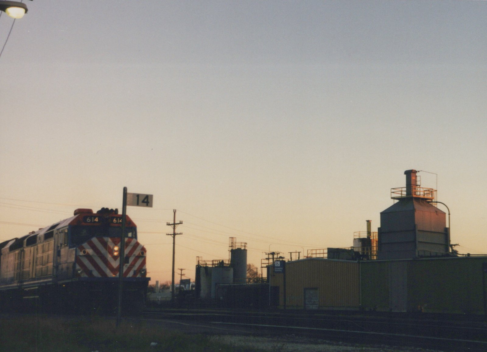A Metra diesel locomotive, number 614, is shown at dusk in a rail yard, its front displaying red and white diagonal stripes, with industrial buildings and a '14' sign in the background.