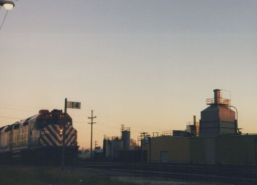 A Metra diesel locomotive, number 614, is shown at dusk in a rail yard, its front displaying red and white diagonal stripes, with industrial buildings and a '14' sign in the background.