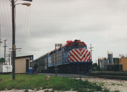 A blue and red Metra locomotive numbered 614 pulls a train of silver passenger cars alongside a grassy station platform under an overcast sky.