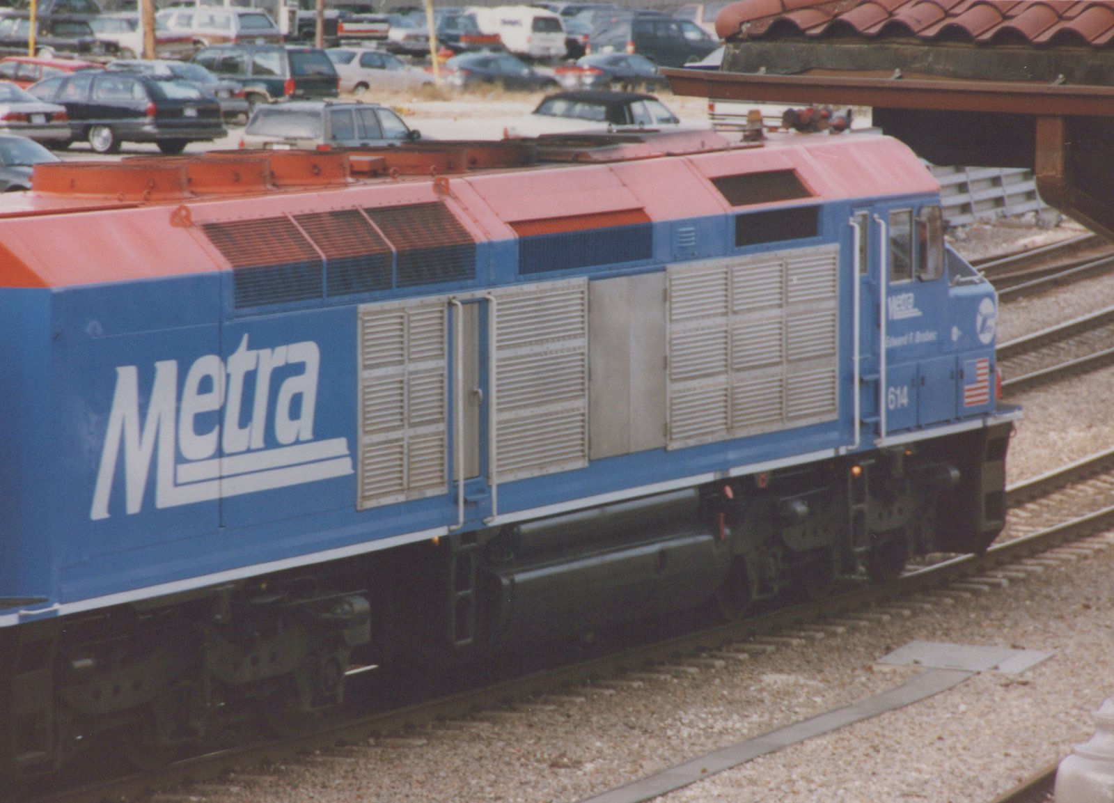A blue and orange Metra diesel locomotive, number 614, named Edward F. Brobst, is shown on tracks beside a station platform with cars parked in the background.