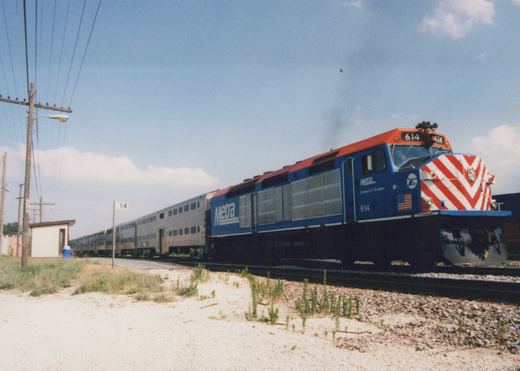 A blue and red Metra diesel locomotive numbered 614 pulls a train of silver bi-level passenger cars on a sunny day, with a small station shelter and utility poles in the background.