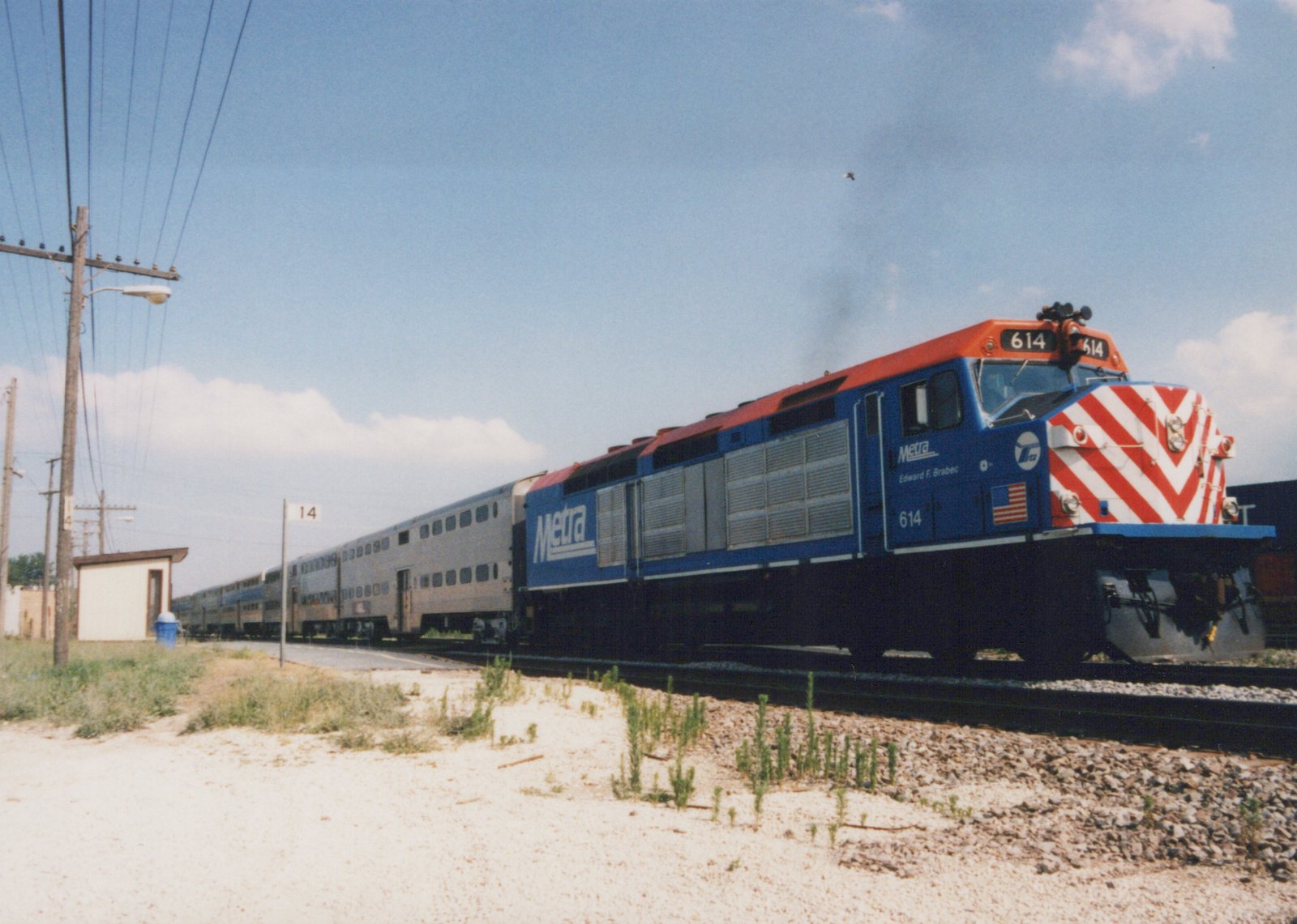 A blue and red Metra diesel locomotive numbered 614 pulls a train of silver bi-level passenger cars on a sunny day, with a small station shelter and utility poles in the background.