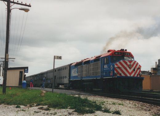 A blue and red Metra diesel locomotive numbered 613 is stopped at a station platform with several silver double-decker commuter cars behind it, under an overcast sky.