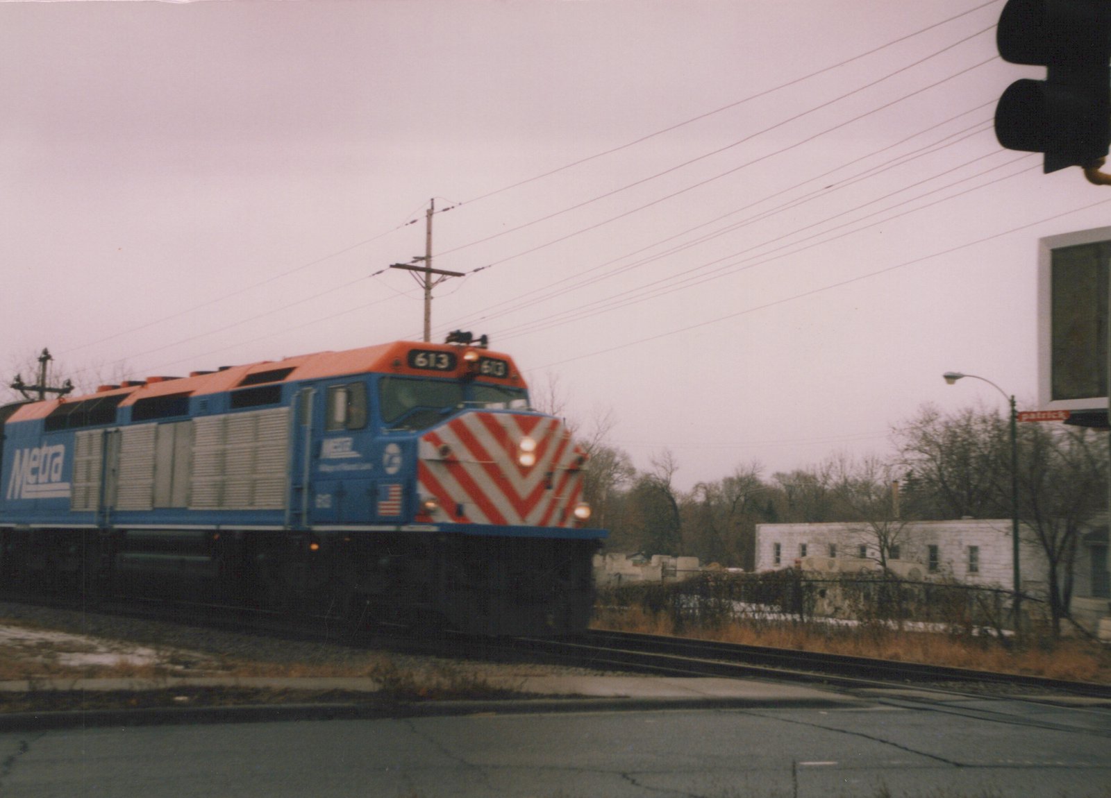 A blue and orange Metra diesel locomotive with the number 613 on its front and side is stopped at a street crossing on an overcast winter day, with bare trees and a traffic light in the background.