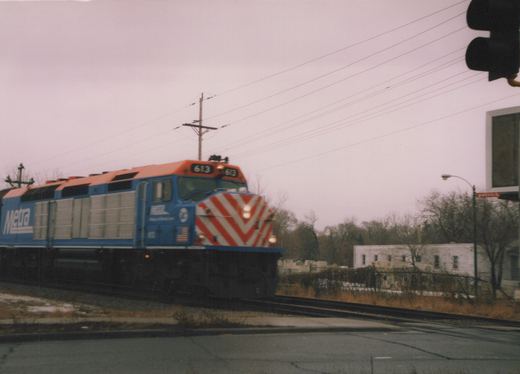 A blue and orange Metra diesel locomotive with the number 613 on its front and side is stopped at a street crossing on an overcast winter day, with bare trees and a traffic light in the background.