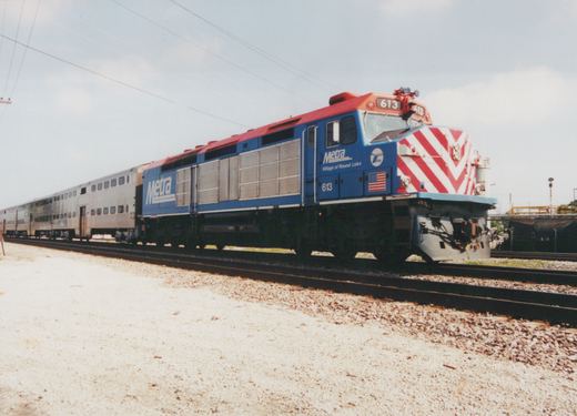 A blue and red Metra locomotive with the number 613 and a tribute to the Village of Round Lake, pulling a line of silver passenger cars on a gravel trackbed under an overcast sky.