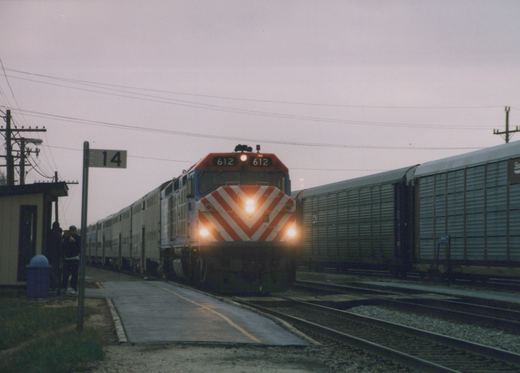 A Metra commuter train with a red and white chevron-nosed locomotive numbered 612 is stopped at a station platform with several people waiting, under an overcast dusk sky.