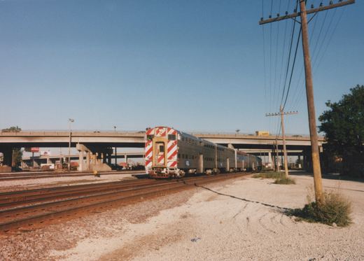 A silver Amtrak railcar with a distinctive red and white striped front end is parked on multiple tracks under a large concrete highway bridge, with utility poles and trees nearby on a sunny day.