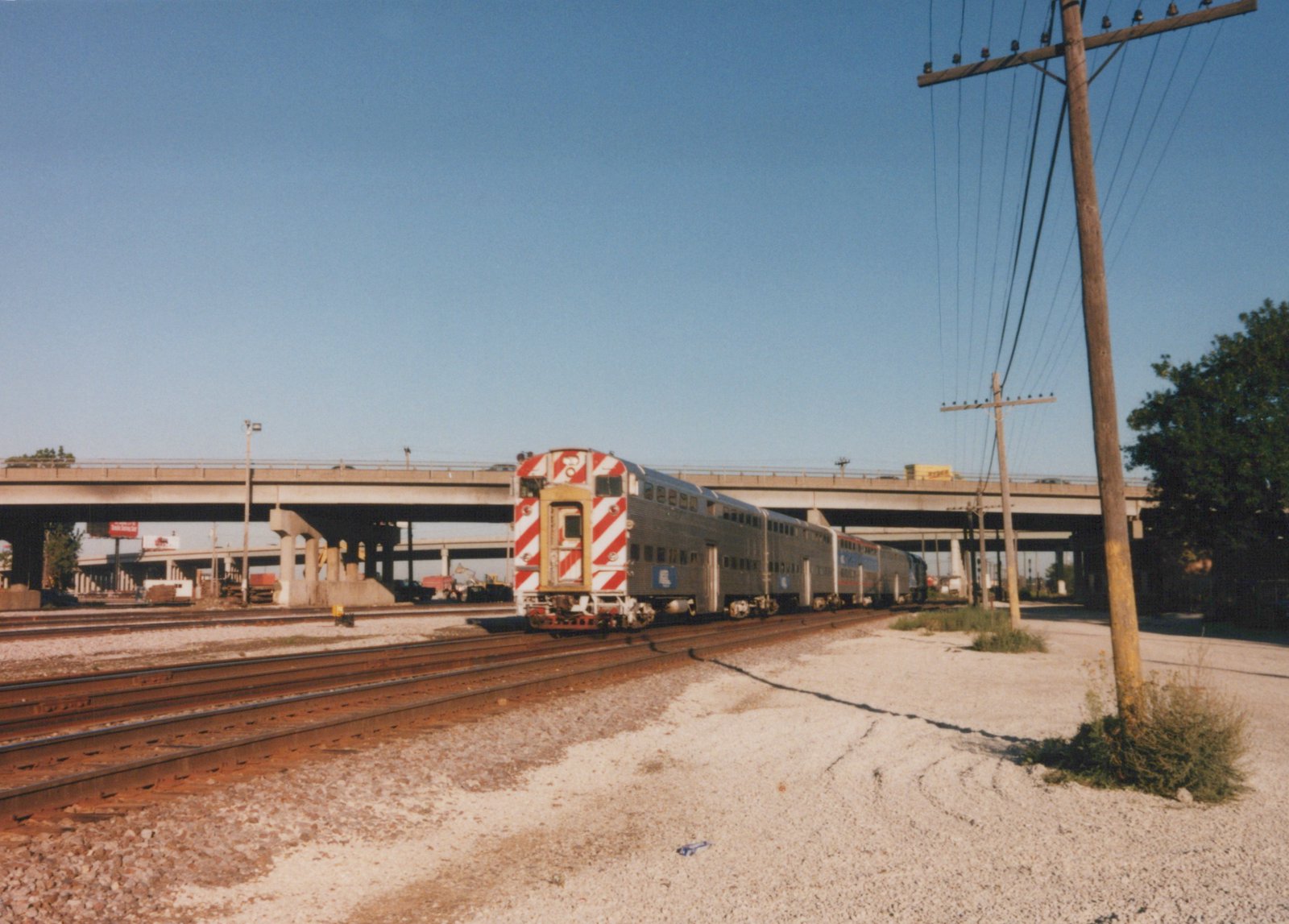 A silver Amtrak railcar with a distinctive red and white striped front end is parked on multiple tracks under a large concrete highway bridge, with utility poles and trees nearby on a sunny day.