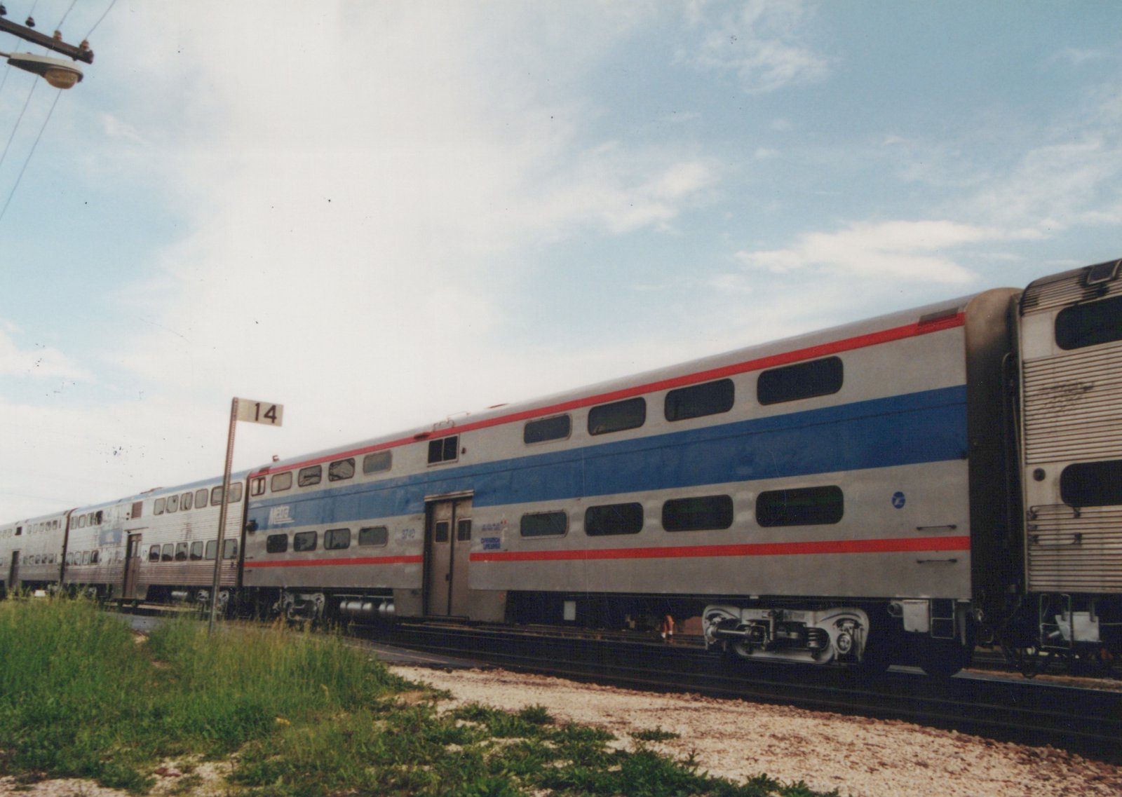 A side view of a Metra Gallery commuter railcar, number 3740, painted silver with blue and red stripes, sitting on tracks next to a grassy area under a partly cloudy sky.