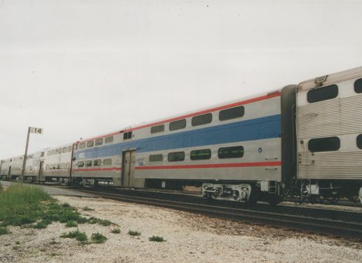 A side view of a silver, blue and red Metra gallery commuter car, numbered 3740, on a set of tracks next to a gravel area with some grass.