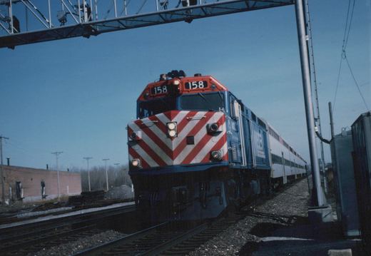A red, white and blue Metra locomotive numbered 158 leads a line of double-decker passenger cars down a set of tracks under a clear winter sky.