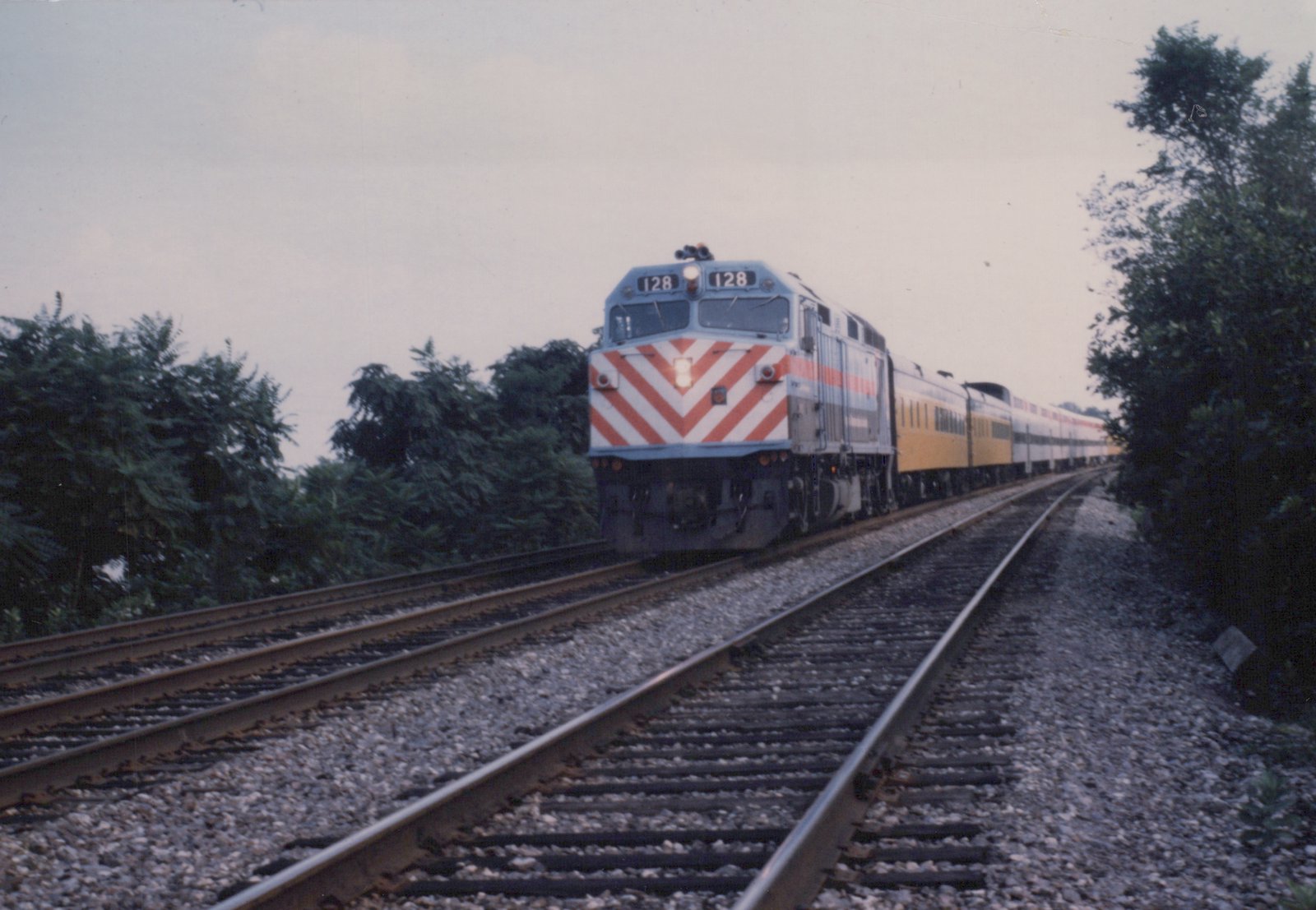 A diesel locomotive with a silver body and red-and-white chevron nose, numbered 128, pulls a line of passenger cars along a gravel-ballasted track flanked by trees under an overcast sky.