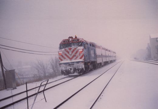 A red, white, and blue Metra locomotive numbered 129 pulls a train of double-decker gallery cars along snow-covered tracks.