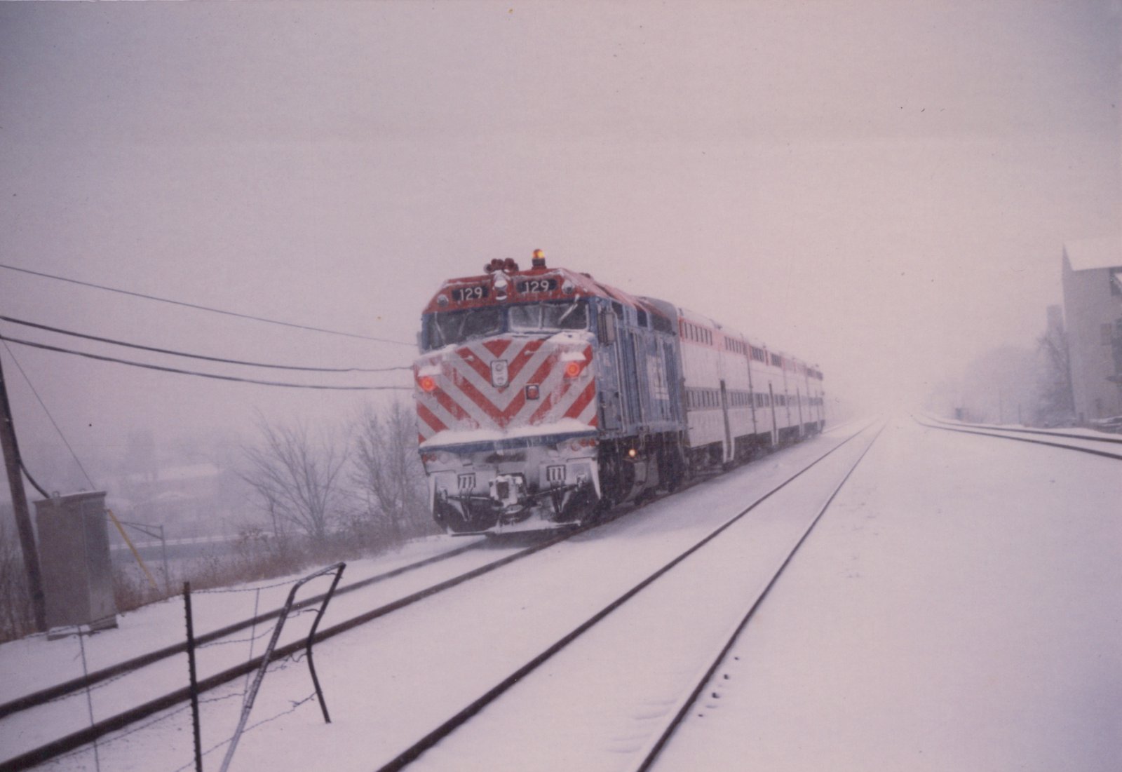 A red, white, and blue Metra locomotive numbered 129 pulls a train of double-decker gallery cars along snow-covered tracks.