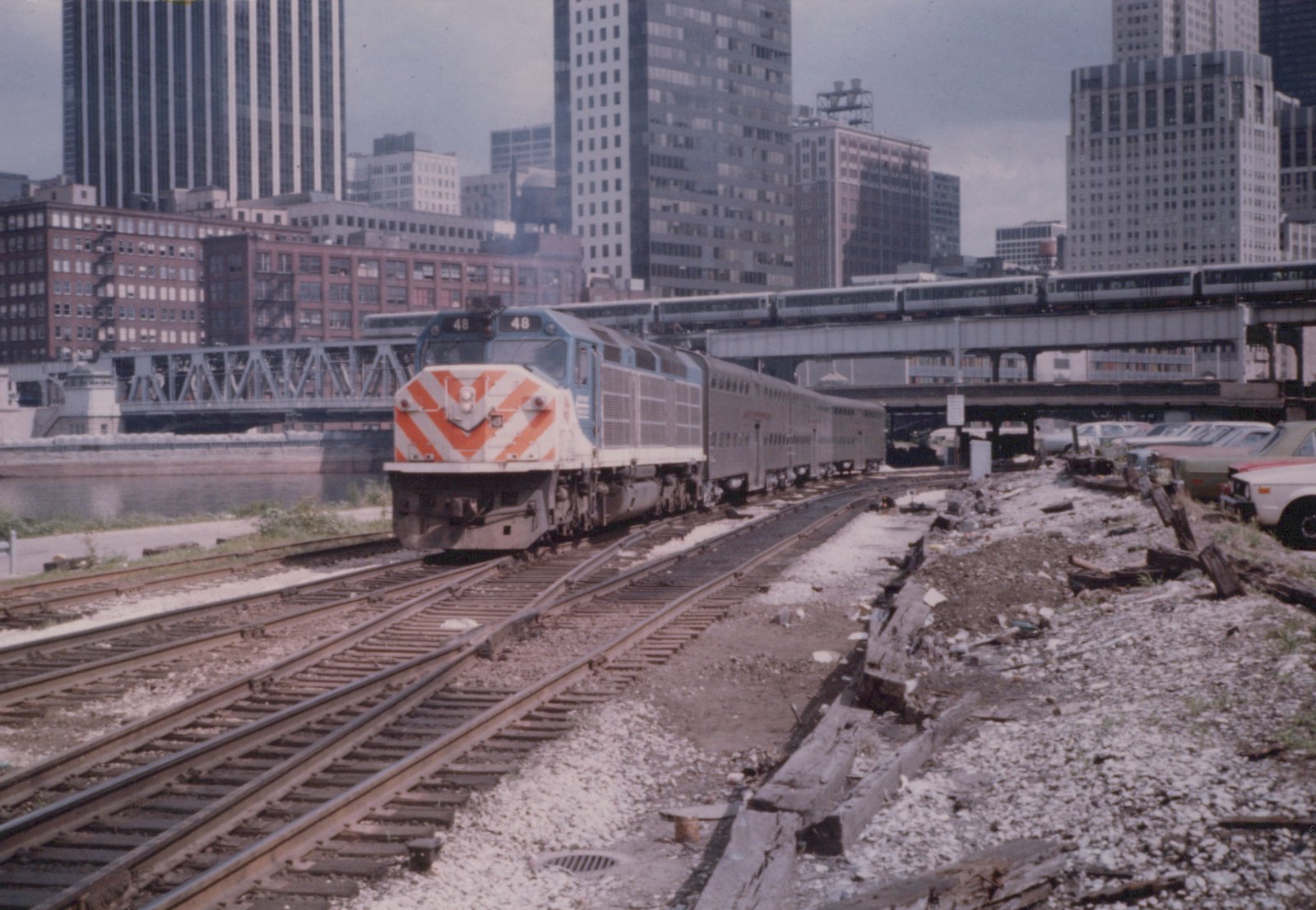 A blue and white diesel locomotive with the number 48 and 'Illinois Central' lettering pulls a train on tracks in a city, with an elevated train and tall buildings in the background.