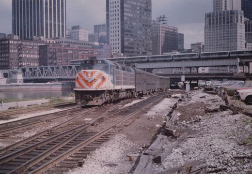 A blue and white diesel locomotive with the number 48 and 'Illinois Central' lettering pulls a train on tracks in a city, with an elevated train and tall buildings in the background.