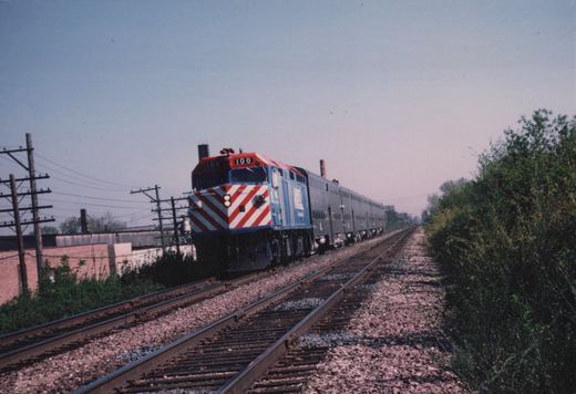 A Metra train, led by a blue and red F40PH locomotive numbered 100, travels along a set of tracks bordered by trees and utility poles on a clear day.