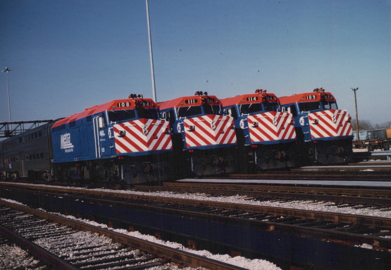 Four blue and red striped Metra diesel locomotives parked side by side in a rail yard, with a gallery car attached to the lead unit, under a clear sky in winter.