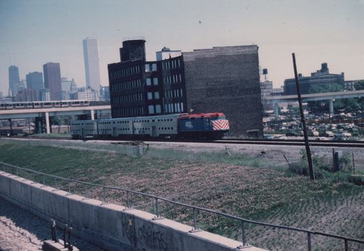 A blue and red Metra locomotive pulls silver passenger cars on tracks beside a grassy slope with graffiti, with city buildings and an elevated train in the background.