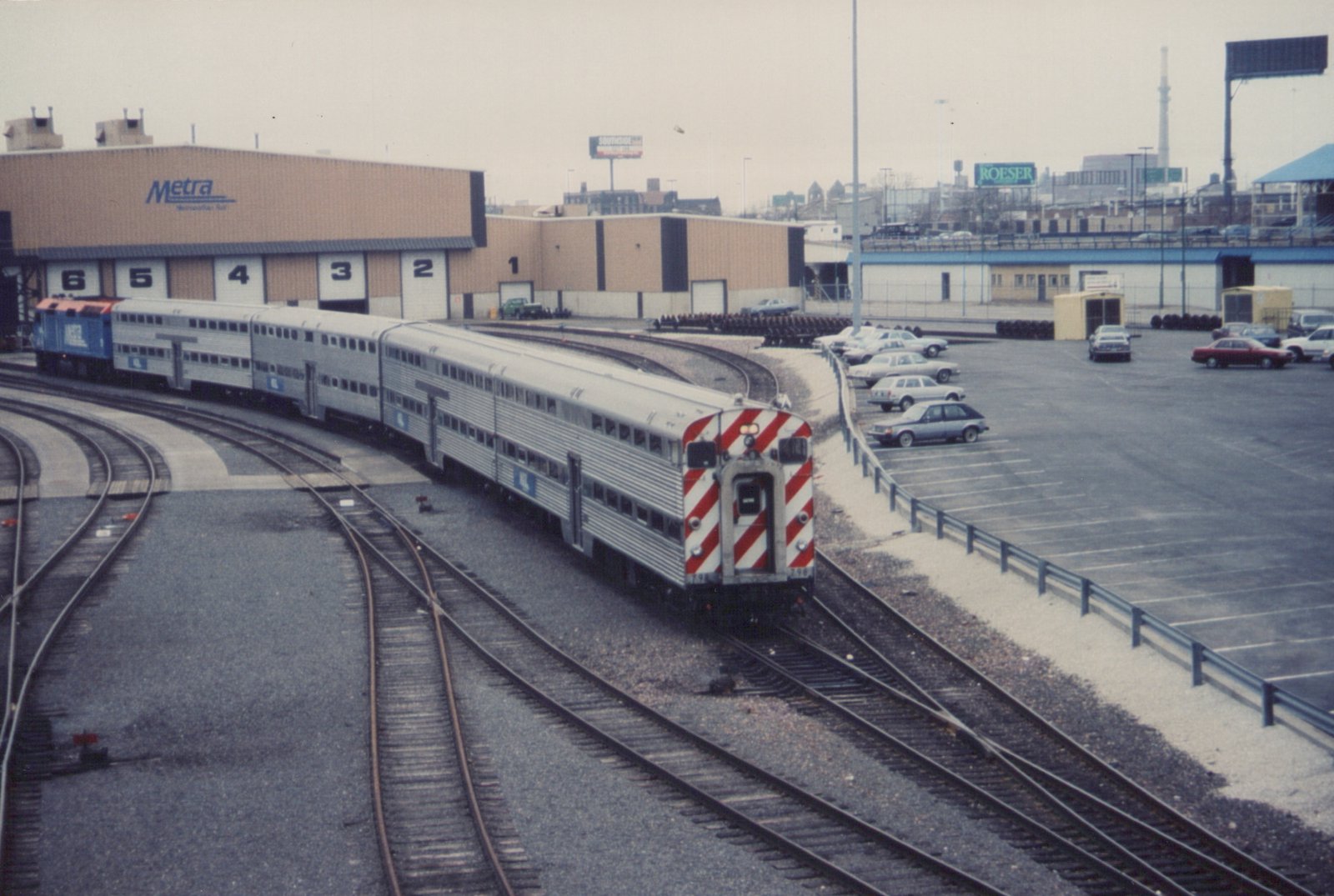 An elevated view of a silver Metra train with red and white striped cab car 398, pulled by a blue F40PH locomotive, curving through a railyard with a large Metra building and a parking lot visible.
