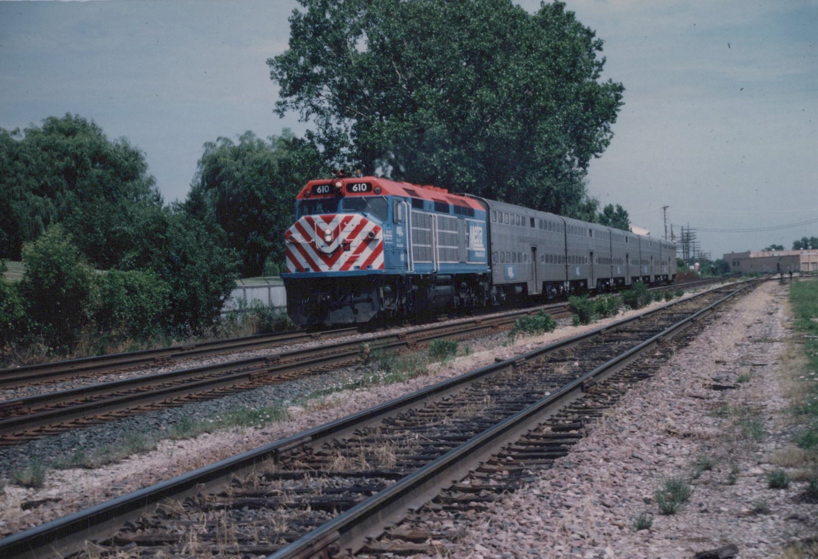A blue and red Metra locomotive numbered 610 leads a train of silver bi-level commuter cars along railroad tracks bordered by green trees under an overcast sky.