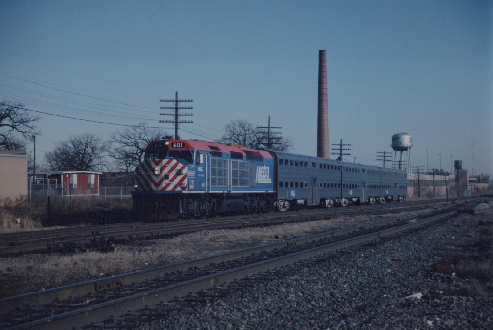 A blue and red Metra locomotive pulling silver bi-level passenger cars along a railroad track with a water tower and smokestack in the background.