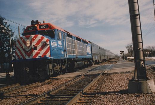 A blue Metra locomotive with a red and white striped nose, numbered 601, pulls a line of stainless steel gallery cars along a station platform on a clear winter day.