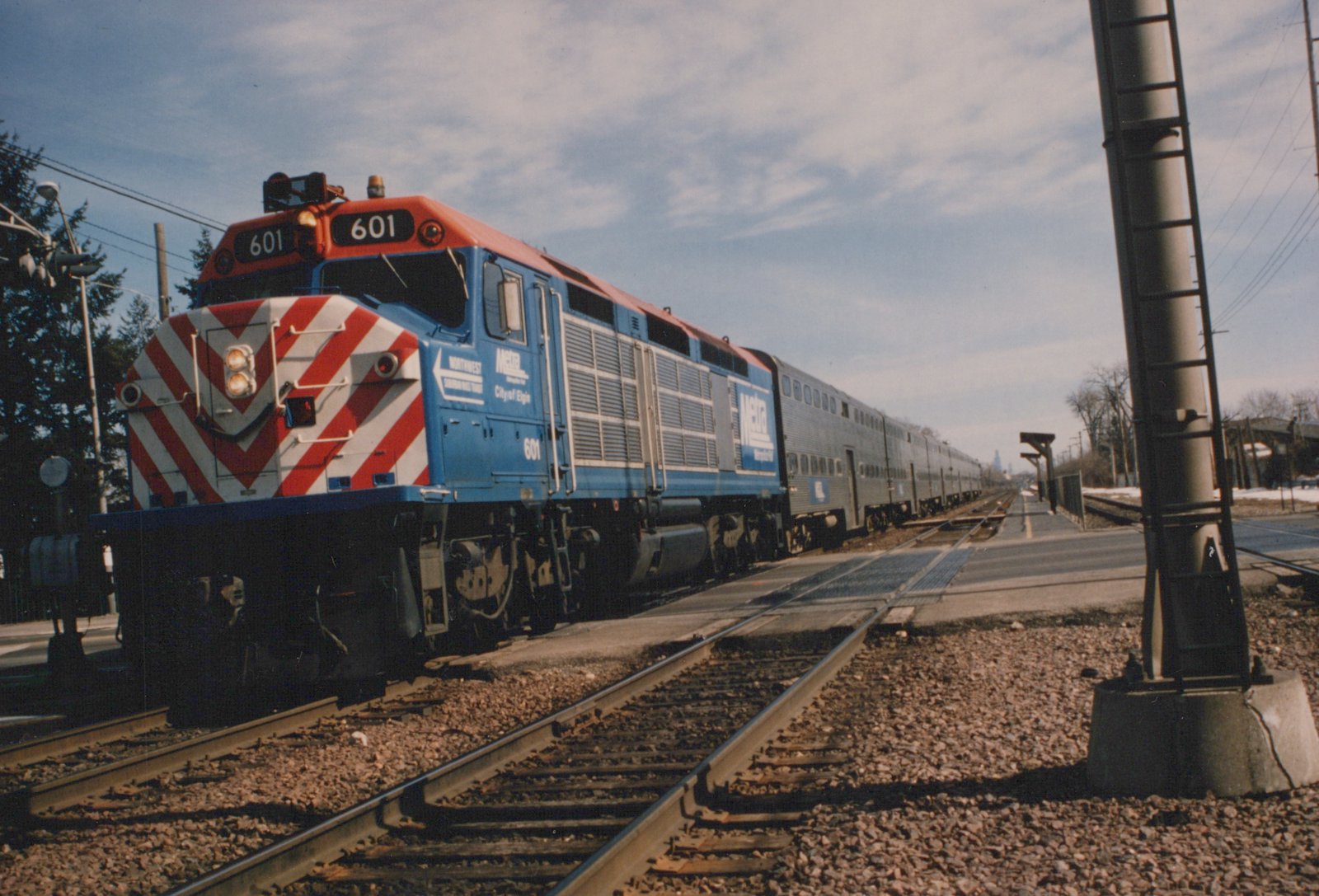 A blue Metra locomotive with a red and white striped nose, numbered 601, pulls a line of stainless steel gallery cars along a station platform on a clear winter day.