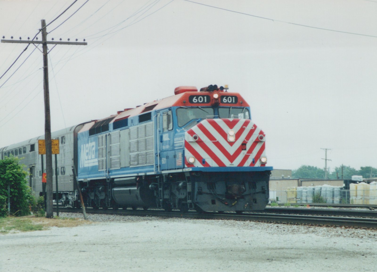 A blue Metra diesel locomotive with number 601 on the front and sides, pulling silver double-decker commuter cars, is shown on railroad tracks next to a utility pole and a yellow railroad crossing sign. The sky is overcast.