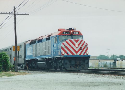 A blue Metra diesel locomotive with number 601 on the front and sides, pulling silver double-decker commuter cars, is shown on railroad tracks next to a utility pole and a yellow railroad crossing sign. The sky is overcast.