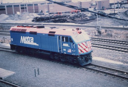 A blue Metra locomotive with a red roof and red-and-white chevron front, number 198, sits on tracks in a gravel yard with a building and maintenance equipment in the background.