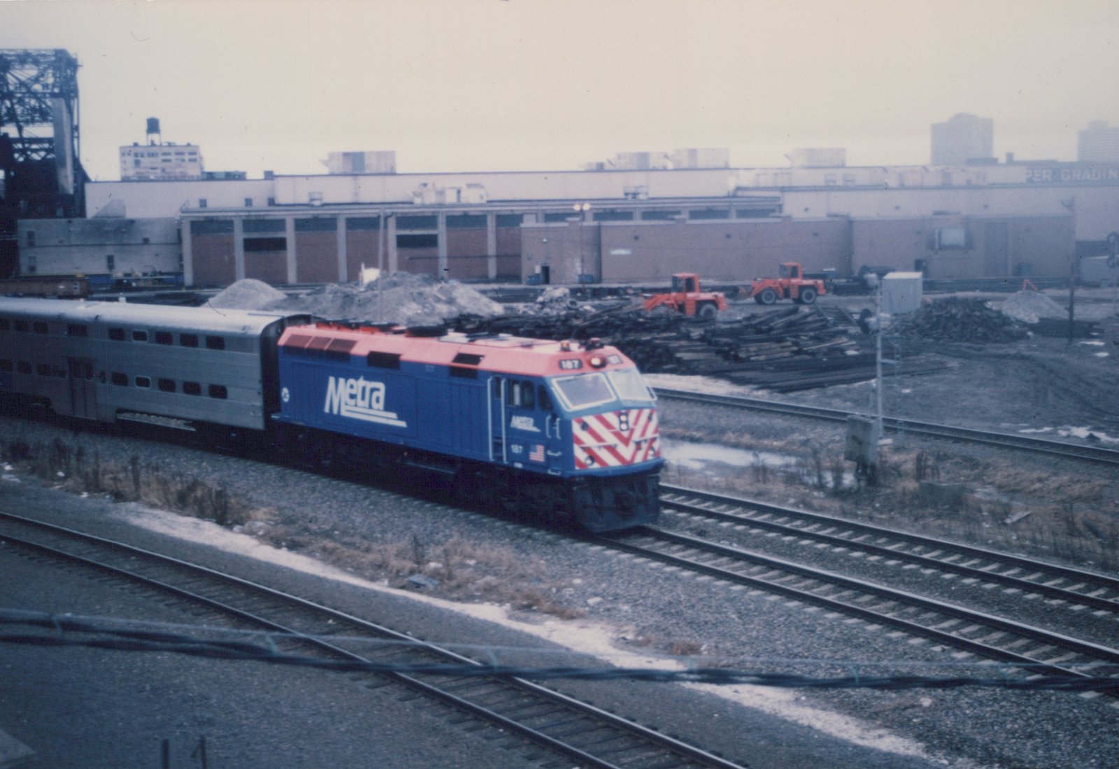 A blue and red Metra locomotive numbered 187 pulls a silver passenger car on multiple tracks in a railyard with an industrial building and orange construction equipment in the background.