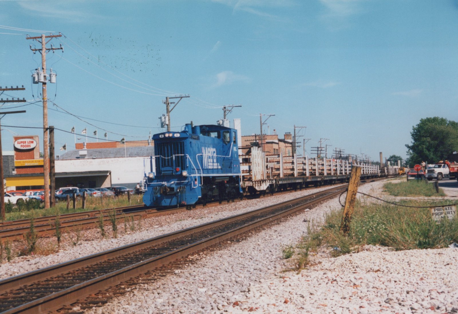 A blue Metra train led by locomotive number 6 hauls a long line of flatcars on a bright, sunny day, with a roadside store and utility poles in the background.