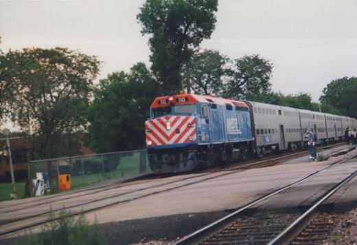 A blue and red Metra locomotive pulls a silver passenger train along railroad tracks next to a platform with people standing on it, set against a backdrop of green trees and an overcast sky.
