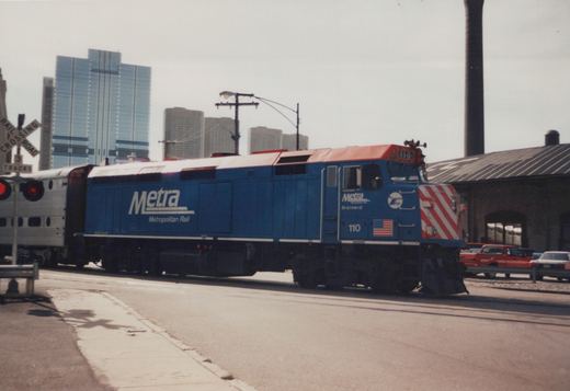 A blue and red Metra diesel locomotive pulls a silver commuter train across a street-level railroad crossing in a city.