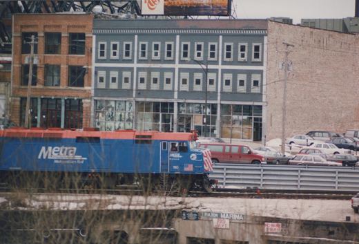 A blue and orange Metra diesel locomotive numbered 110 is shown on an elevated track with snow on the ground, buildings in the background, and a sign reading 'Fulton Marina' below.