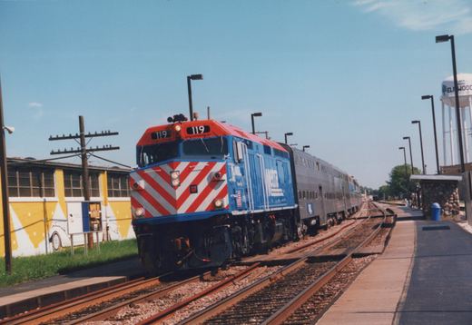 A blue and red Metra locomotive with the number 119 on its front pulls a silver train of bi-level coaches along tracks next to a station platform, with a water tower labeled 'ELMWOOD' in the background under a clear blue sky.
