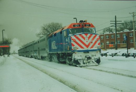 A blue and red Metra locomotive numbered 121 pulls a line of silver double-decker passenger cars along snow-covered tracks, with snow-covered cars and a brick building in the background.