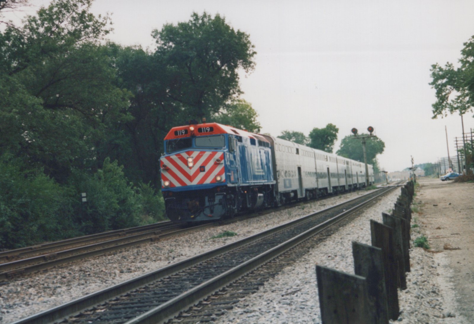 A red, white, and blue Amtrak train with a locomotive numbered 119 pulls several silver passenger cars along a set of tracks bordered by trees and a wooden fence.