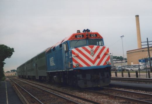 A blue Metra locomotive with a red and white striped front, numbered 125, is pulling a line of double-decker passenger cars on railroad tracks next to a station platform.