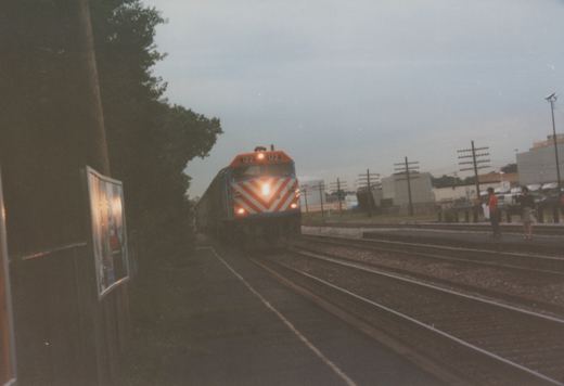 A Metra diesel locomotive with number 122 on its front, painted in orange and white chevrons, moves toward the camera on a set of tracks with two people waiting on a platform to the right.