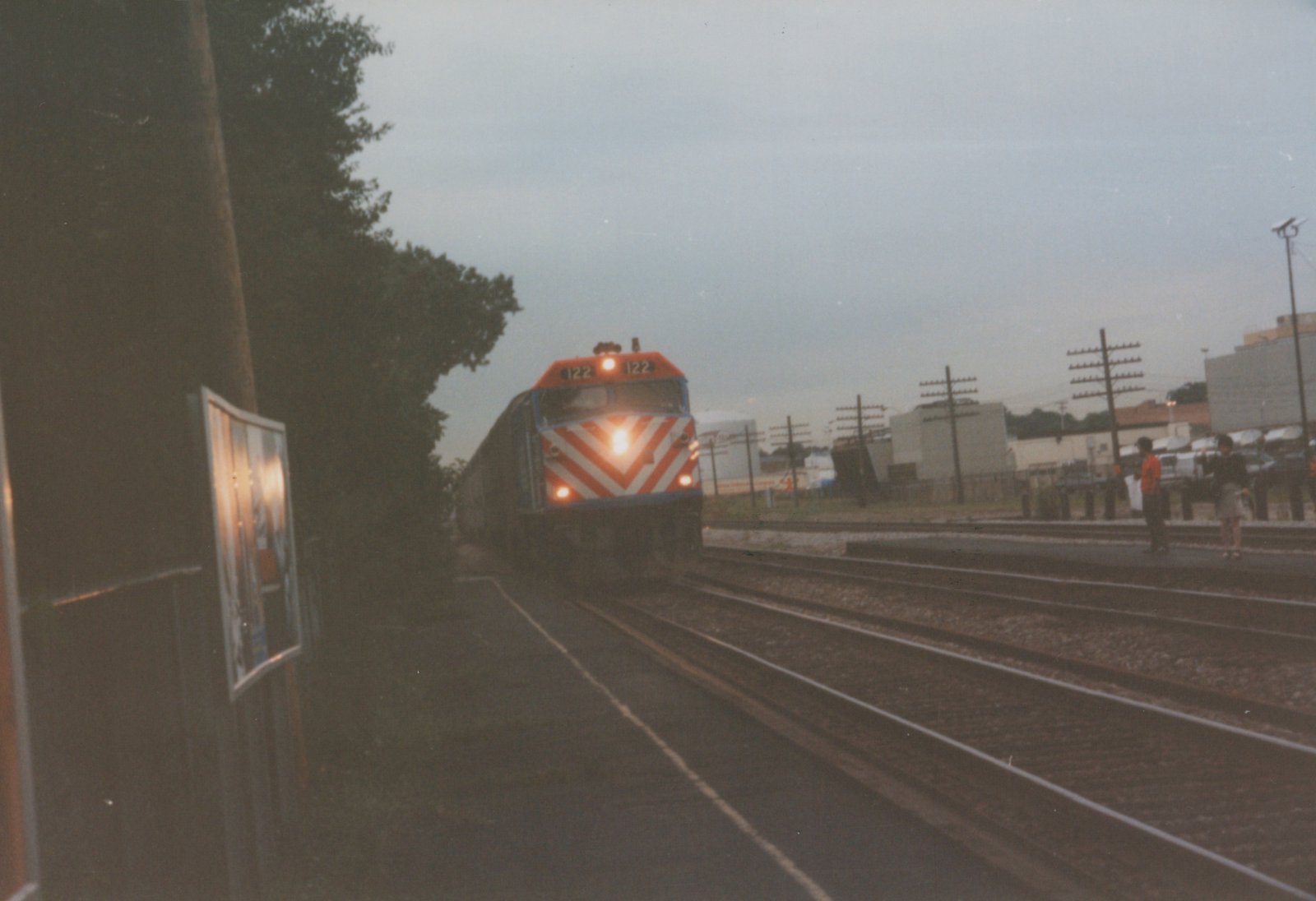 A Metra diesel locomotive with number 122 on its front, painted in orange and white chevrons, moves toward the camera on a set of tracks with two people waiting on a platform to the right.